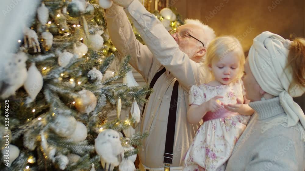 Elderly man decorating Christmas tree at home as his wife holding adorable toddler granddaughter on arms Elderly man decorating Christmas tree at home as his wife holding adorable toddler granddaughter on arms