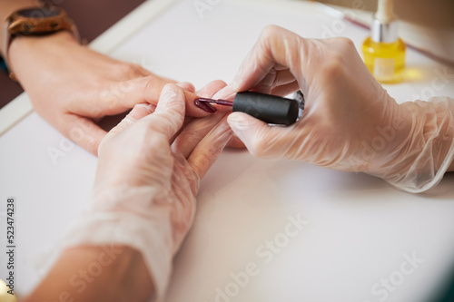 Professional manicurist doing manicure for woman client in beauty salon. Close up of manicure specialist hands in sterile gloves applying red nail polish on woman fingernail.