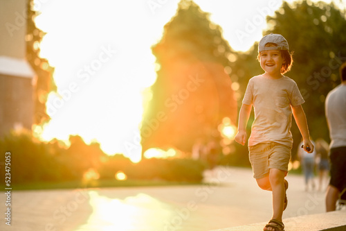 A cute boy with long hair and a baseball cap walks in the summer in the park against the backdrop of the setting sun