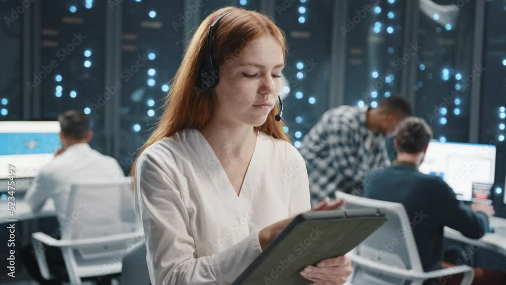 Close up cute young smiling Caucasian redhead woman in headset wearing formal clothes, holding tablet in hands standing in center of open space of office speaking with client Close up cute young smiling Caucasian redhead woman in headset wearing formal clothes, holding tablet in hands standing in center of open space of office speaking with client