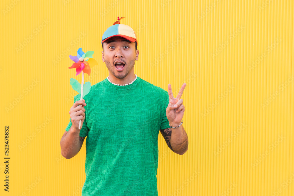 Young man with rainbow pinwheel toy showing peace sign in front of wall Young man with rainbow pinwheel toy showing peace sign in front of wall