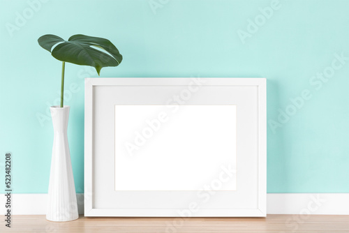 Elegant horizontal transparent white picture frame with matte and monstera leaf in vase in front of pastel green wall.