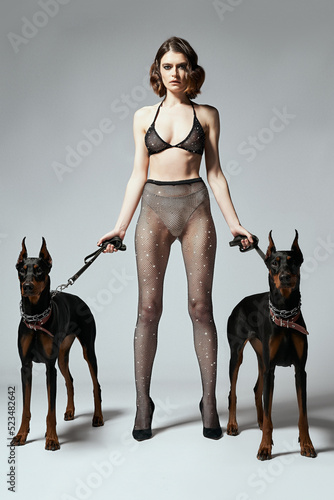 Close-up shot of a young woman keeping two dogs on leads. The girl in black transparent tights and a bra decorated with a scattering of sparkling crystals is on a gray background. Front view.