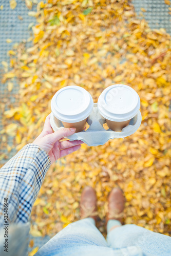 Woman hands holding two cups of takeaway coffee on autumn yellow leaves background with copy space