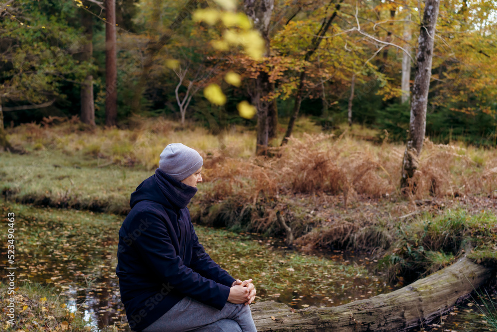 Young man in warm clothes sitting on a fallen tree near river, looking at the forest and enjoying the moment. Feeling harmony, reunion with nature in autumn. Relaxing, personal fulfillment