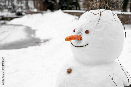 Old funny fat snowman with nose carrot nose, buttons made of walnuts, cork eyes stands alone in open snow-covered field near frozen lake in park on quiet winter december, january or february day