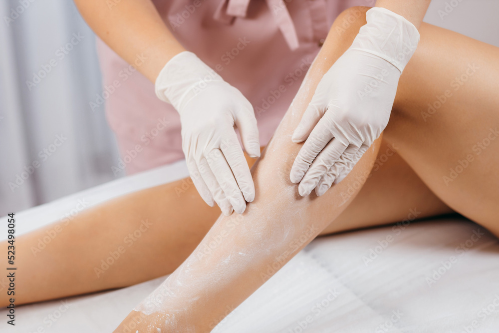 Naklejka premium Hands of beautician in gloves apply cosmetic talcum on legs of women model powder before 