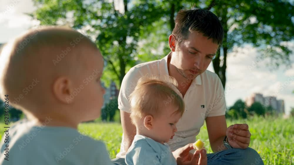 Father gives baby son small piece of banana sitting on picnic grass. Baby boy examines food and takes from father hand. Child looks with serious expression closeup Father gives baby son small piece of banana sitting on picnic grass. Baby boy examines food and takes from father hand. Child looks with serious expression closeup