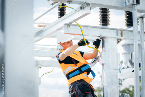 Electrician in protective helmet working on high voltage power lines. Highly skilled workmen servicing the electricity grid. Modern power station with power towers