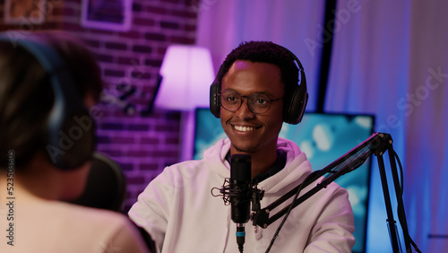 Portrait of smiling african american online radio host listening to interesting conversation with influencer in home recording studio. Podcaster broadcasting live with guest using microphone. © DC Studio