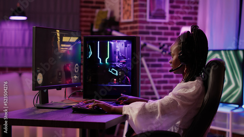 African american woman streaming first person shooter while talking in headset in home living room. Gamer girl streamer using pc gaming setup having a good time playing multiplayer online action game