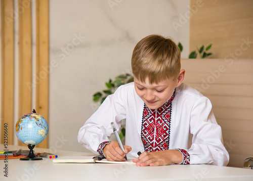schoolboy in ukrainian embroidery writing in notebook and sitting at the desk at school with globe and different colored penciles