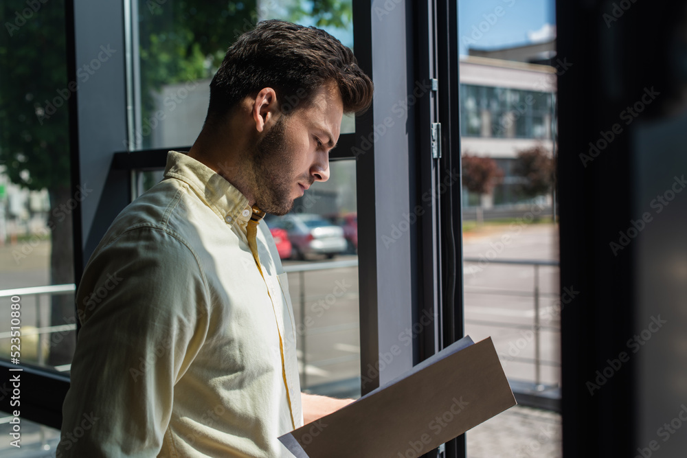 Side view of businessman looking at paper folder near window in office. Side view of businessman looking at paper folder near window in office.