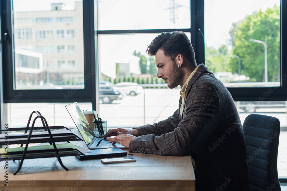 Side view of businessman in earphone using laptop near paperwork in office. Side view of businessman in earphone using laptop near paperwork in office.