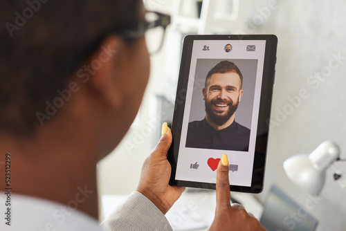 Young woman finds her love online. Happy African American girl gives like to picture of smiling handsome Caucasian man on modern mobile love app or dating website. Close up of hands and tablet screen photo