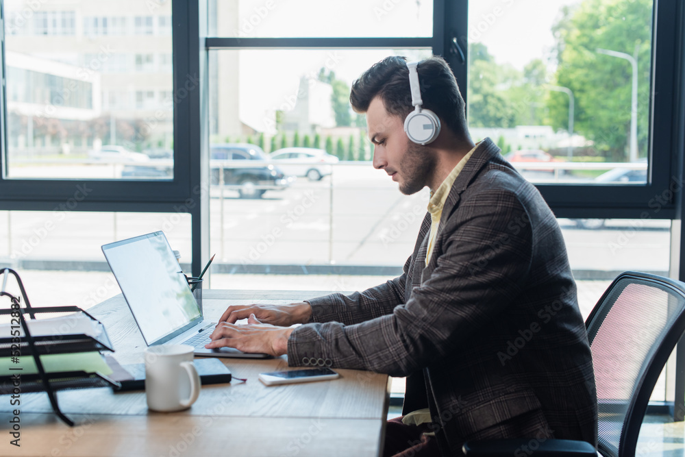 Side view of young businessman in headphones using laptop near paperwork and cup in office.