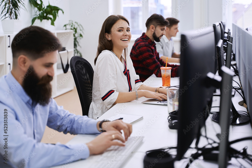 Junior female professional working in modern IT office. Group of young and experienced programmers and software developers sitting at desks working on computers.