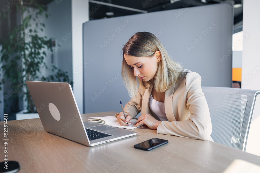 Fototapeta premium Serious women in classic clothes, writing notes about the training for students, works at the table with a laptop.