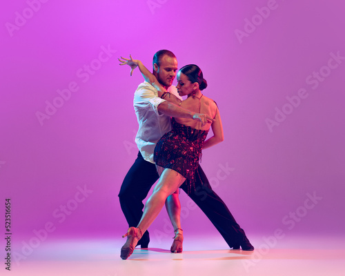 Two young graceful dancers wearing stage outfits dancing ballroom dance isolated on purple background. Concept of art, beauty, music, style.