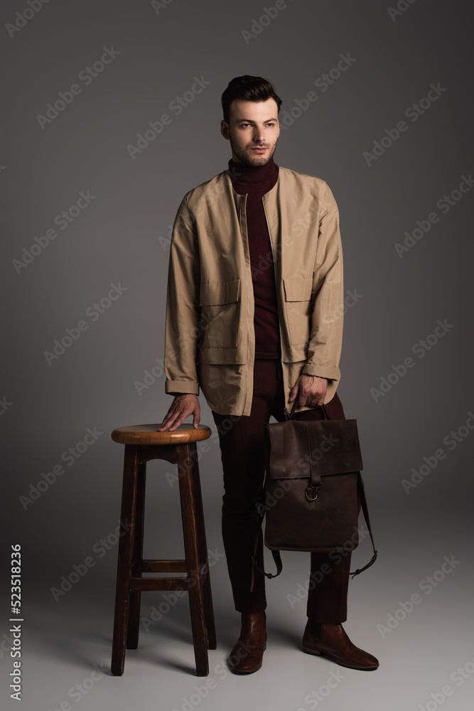 Full length of bearded man in autumn outfit holding backpack near chair on grey background. Full length of bearded man in autumn outfit holding backpack near chair on grey background.