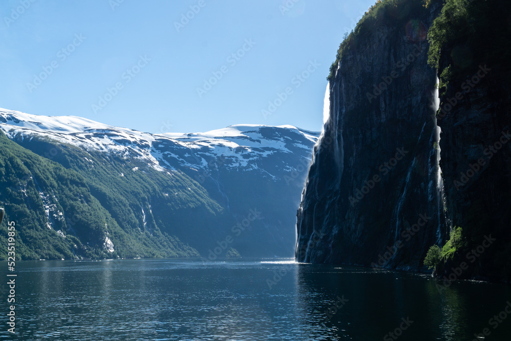 Famous seven sisters waterfall sliding down the cliff into Geirangerfjord on a sunny day with snow-capped mountains in the background.