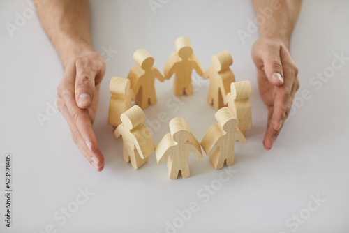 Human hands protecting and guarding small wooden toy figures placed on white desk as metaphor for creating safe, supportive community of people. Close up, closeup shot. Care, support, safety concepts