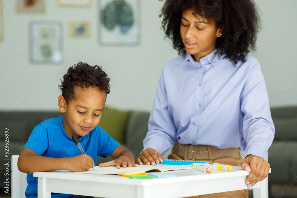 Female infant school teacher working one on one with a young schoolboy, sitting at a table writing in a classroom, front view Female infant school teacher working one on one with a young schoolboy, sitting at a table writing in a classroom, front view