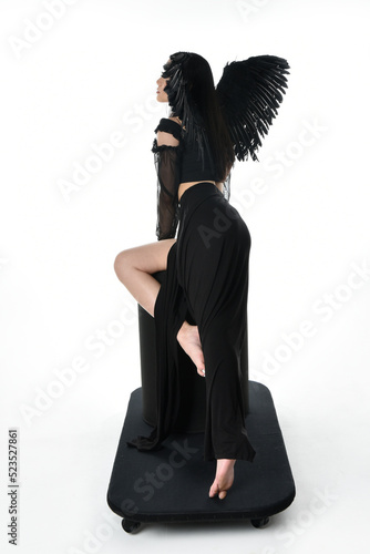 full length portrait of beautiful asian model with dark hair, wearing black gothic skirt costume, angel feather wings with horned headdress. crouching pose isolated on studio background.