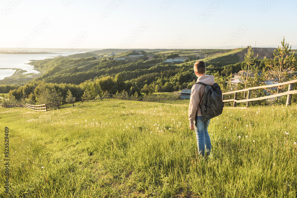 Tourist with backpack standing on top of hill in grass field and enjoying beautiful landscape view. Rear view of teenage boy hiker resting in nature. Active lifestyle. Concept of local travel Tourist with backpack standing on top of hill in grass field and enjoying beautiful landscape view. Rear view of teenage boy hiker resting in nature. Active lifestyle. Concept of local travel