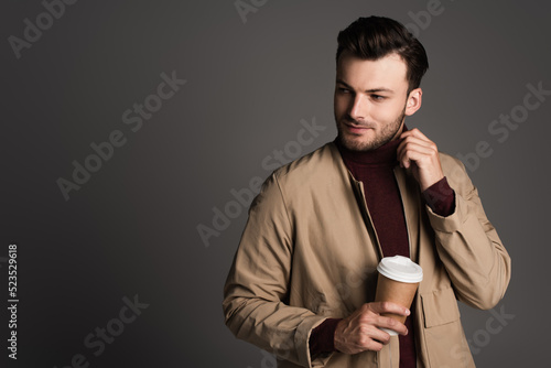 Fashionable young man in autumn jacket holding takeaway coffee isolated on grey.