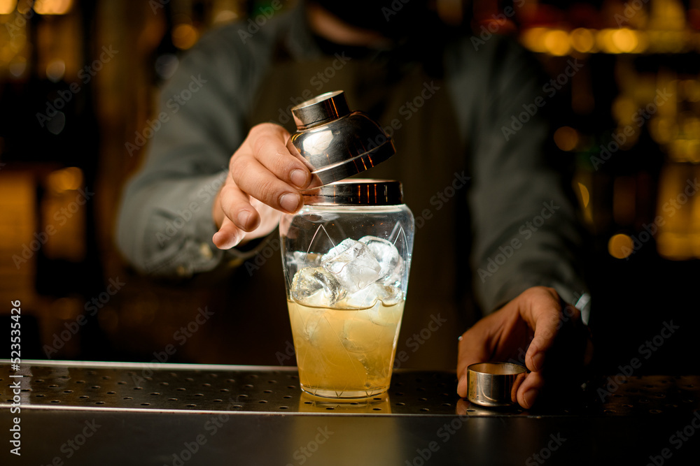 close-up of transparent glass shaker with liquid a which the hand of bartender closes with a lid