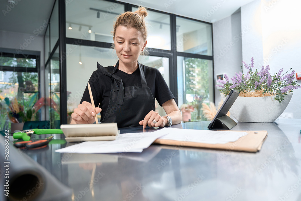 Happy florist is sitting at table and counting on calculator