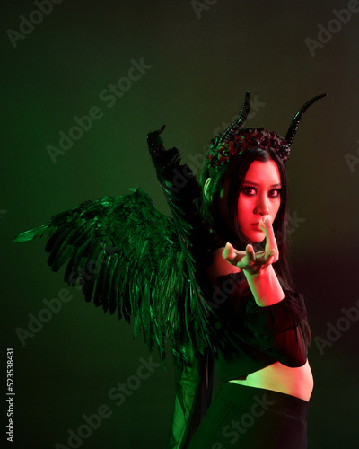 portrait of beautiful asian model with dark hair, wearing black gothic skirt costume, angel feather wings with horned headdress. Posing with gestural hands on dark silhouette studio background.