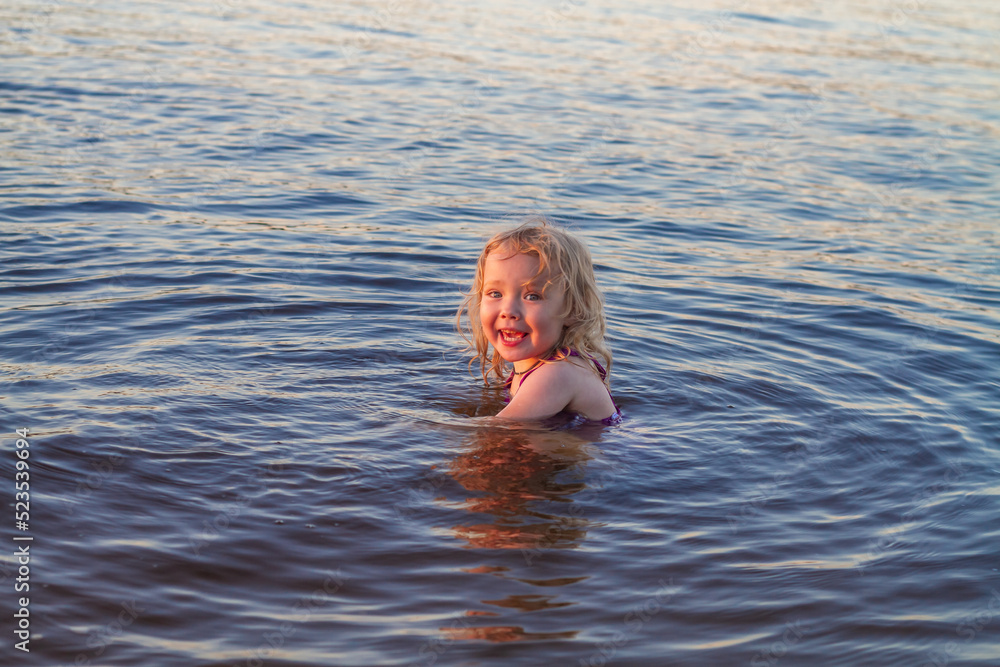 Gentle baby blonde girl 3 years old swims in the water in the river, lake, sea in summer in a purple swimsuit