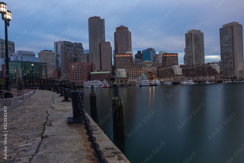 Boston Cityscape With A Walkway And Twin lit street lamp lanterns On The Side, Boats And Cruise Ships Anchoring Near The Still Water Front Under Blue Cloudy Sky
