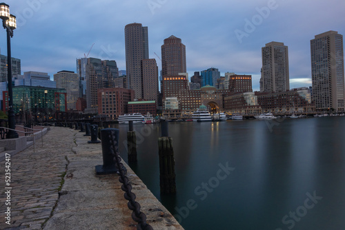 Boston Cityscape With A Walkway And Twin lit street lamp lanterns On The Side, Boats And Cruise Ships Anchoring Near The Still Water Front Under Blue Cloudy Sky © Gentle.Cam