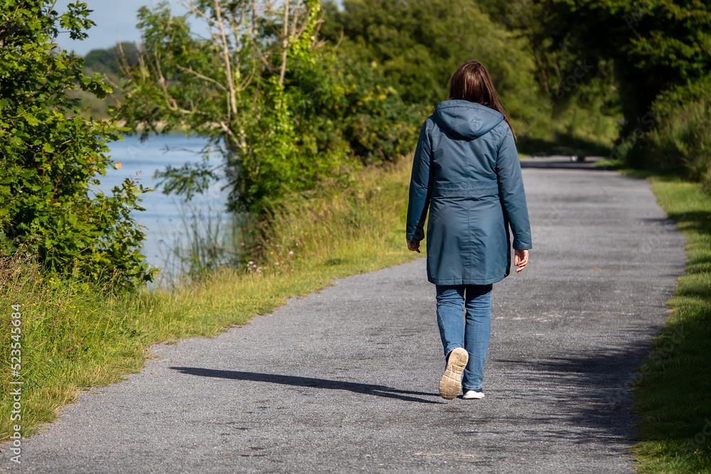Woman walking on a footpath in a park by a river. Back to camera. Warm sunny day. Outdoor activity. Woman walking on a footpath in a park by a river. Back to camera. Warm sunny day. Outdoor activity.