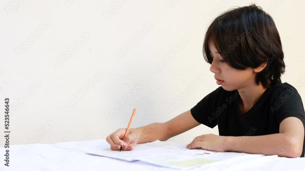 Young east asian teenager boy is studying writing with a pen and notebook at the table. The boy who does his school homework at home. Black tshirt on isolated white background Young east asian teenager boy is studying writing with a pen and notebook at the table. The boy who does his school homework at home. Black tshirt on isolated white background
