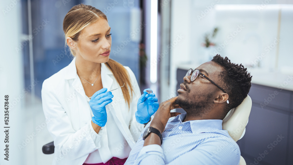 Unhappy african american man in medical chair complains of toothache to female dentist at modern dental clinic. Shot of a young man having a consultation with his dentist Unhappy african american man in medical chair complains of toothache to female dentist at modern dental clinic. Shot of a young man having a consultation with his dentist