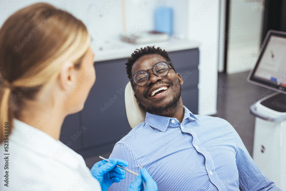 Fototapeta premium Female dentist with happy male patient at clinic. Happy patient sitting on chair while looking at dentist in medical clinic. Cheerful african american man with during examination in dental clinic