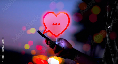 illustrierte rote Sprechblase in Form eines Herzens über einer Hand, die ein Smartphone mit Bildschirm hält, der in einer romantischen Nachtatmosphäre leuchtet, Lichter,, Dating, Flirt, Liebe