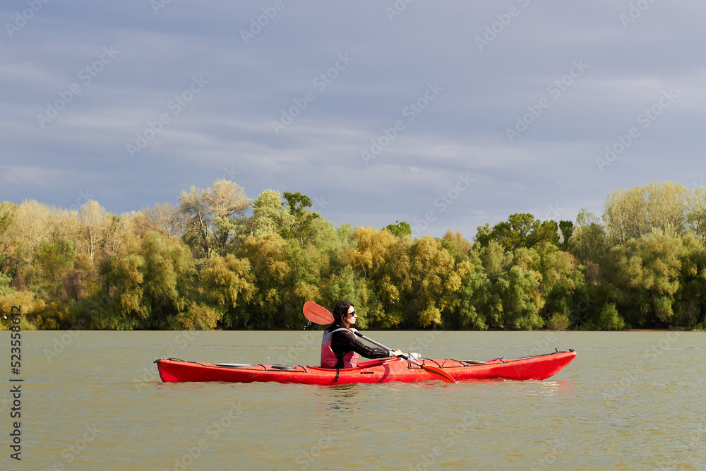 A girl on a red kayak paddles along the Danube river in spring against dark blue sky and gently green trees. Ukraine