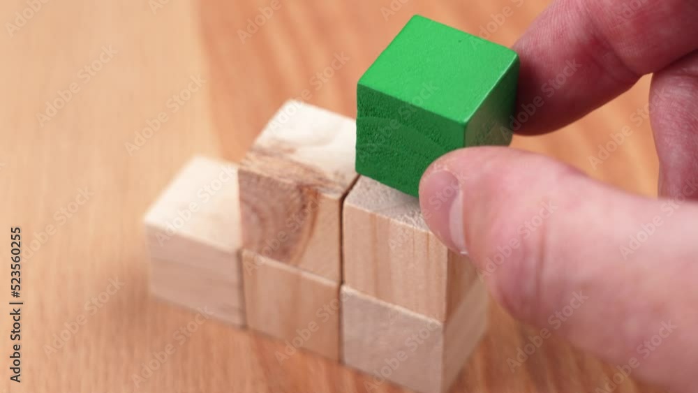 A hand places a bright green wooden cube on top of a wood block tower. Sustainable environment and eco industries business concept A hand places a bright green wooden cube on top of a wood block tower. Sustainable environment and eco industries business concept