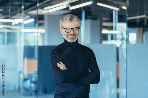 Portrait of successful businessman in modern office with crossed arms, gray haired male investor smiling and looking at camera, boss in glasses and jeans