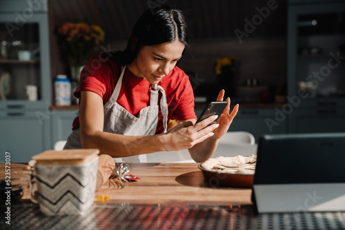 Young woman wearing apron taking photo of meal while cooking at home