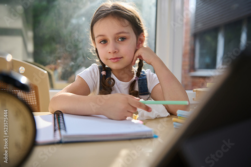 Portrait of beautiful European little girl, primary student, schoolgirl with two pigtails, confidently looking at camera while sitting at desk, learning the writing while studying in an online school.