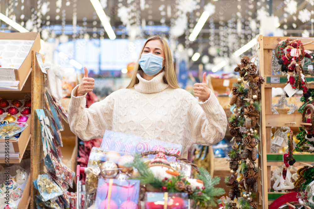 Christmas shopping. Woman with cart shopping in protective medical mask watching Christmas ornament in shopping mall on Holiday sale.