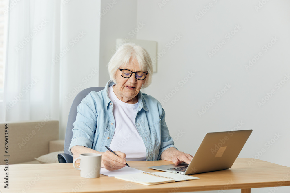 a pleasant, stylish elderly woman is sitting at home in a bright interior and working on a laptop makes notes on a piece of paper. The concept of working from home a pleasant, stylish elderly woman is sitting at home in a bright interior and working on a laptop makes notes on a piece of paper. The concept of working from home