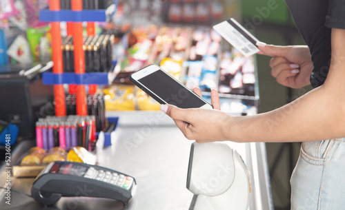 Caucasian woman using smartphone and holding credit card in supermarket.