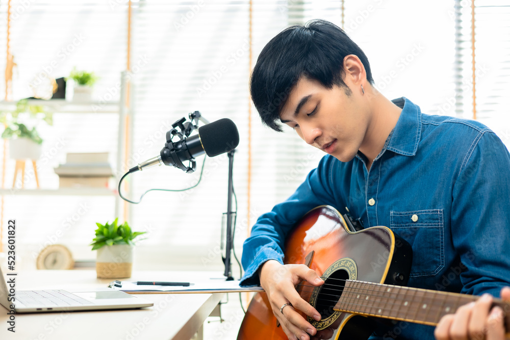 Asian young man songwriter play acoustic guitar listen song from smartphone think and write notes lyrics song in paper sit in living room at home studio. Music production at home concept.